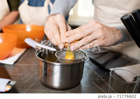 Hands of senior man in apron breaking egg into sieve over metallic pan by table 71776602
