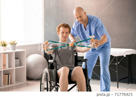 Mature clinician in blue uniform supporting hand of young patient in wheelchair Mature clinician in blue uniform supporting hand of young patient in wheelchair 71776925