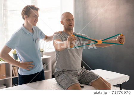 Bald man stretching resistance band during exercise for arms on medical couch Bald man stretching resistance band during exercise for arms on medical couch 71776970