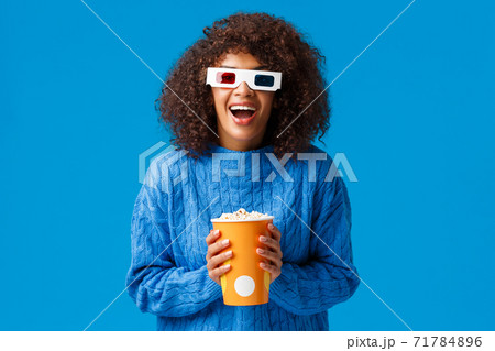 Cheerful gorgeous african-american woman enjoying watching movie in cinema, laughing and smiling excited with 3d effects wear glasses, holding popcorn, standing blue background 71784896