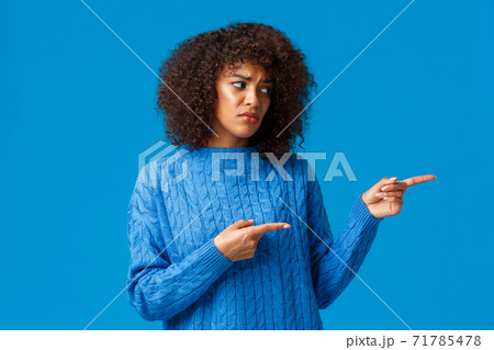 Unhappy sad and timid, insecure lovely african-american pretty woman with afro haircut in winter sweater, sighing uneasy looking pointing right with jealousy or regret, standing blue background Unhappy sad and timid, insecure lovely african-american pretty woman with afro haircut in winter sweater, sighing uneasy looking pointing right with jealousy or regret, standing blue background 71785478