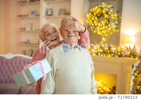 Elderly woman giving christmas gift to her husband and closing his eyes with her hand Elderly woman giving christmas gift to her husband and closing his eyes with her hand 71790223