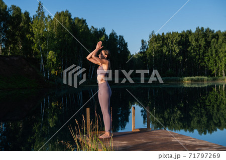 Young woman by the lake practicing yoga moves on wooden platform. Pretty young woman exercising in nature, healthy lifestyle young people positive vibes. 71797269