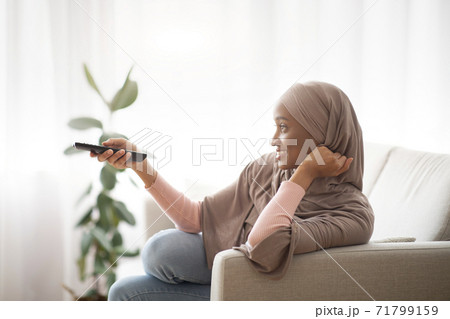 Side view of lovely black woman in traditional hijab holding remote control and watching TV indoors, side view Side view of lovely black woman in traditional hijab holding remote control and watching TV indoors, side view 71799159