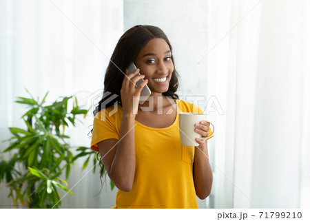 Millennial black woman speaking on cellphone and drinking coffee near window at home 71799210
