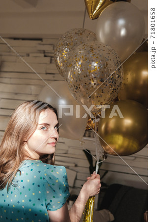 Young beautiful girl in blue dress with white polka dots celebrates her birthday and enjoys the golden balloons. Birthday alone at home during self-isolation. Young beautiful girl in blue dress with white polka dots celebrates her birthday and enjoys the golden balloons. Birthday alone at home during self-isolation. 71801658