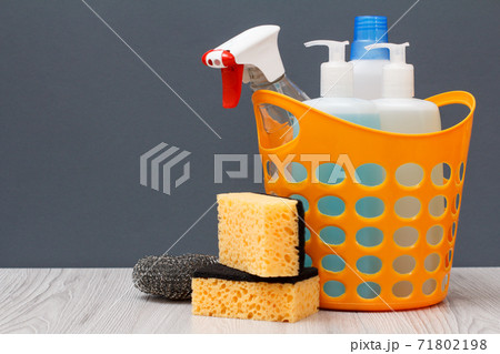 Bottles of dishwashing liquid and sponges in a basket on wooden desk. Bottles of dishwashing liquid and sponges in a basket on wooden desk. 71802198