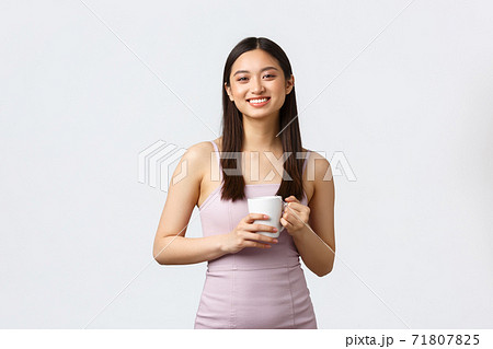 Lifestyle, people emotions and glamour concept. Gorgeous young korean girl in stylish dress, drinking coffee and smiling as talking to coworker, holding mug, standing white background 71807825