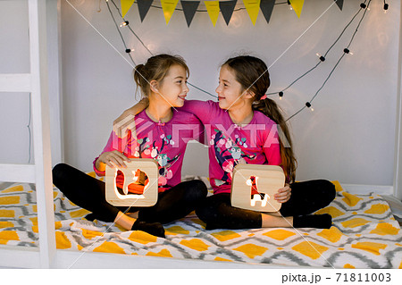 Happy children, two cute 10 year old girls sisters, in child's room on a bunk-bed, sitting in lotus position, looking each other and holding wooden night lamps with cut out pictures. 71811003