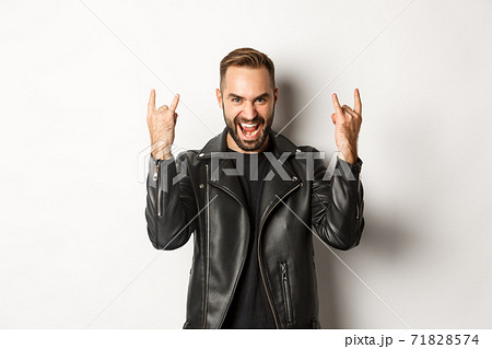 Cool adult man in black leather jacket, showing rock on gesture and tongue, enjoying music festival, standing over white background 71828574