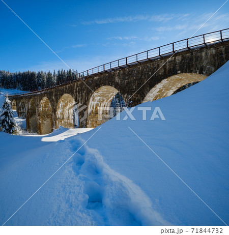 Stone viaduct (arch bridge) on railway through mountain snowy fir forest. Snow drifts on wayside and hoarfrost on trees. Stone viaduct (arch bridge) on railway through mountain snowy fir forest. Snow drifts on wayside and hoarfrost on trees. 71844732