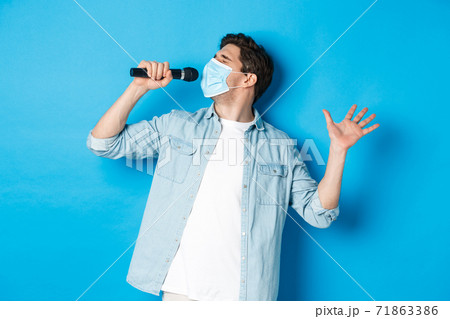 Handsome guy in medical mask singing in microphone, standing over blue background Handsome guy in medical mask singing in microphone, standing over blue background 71863386