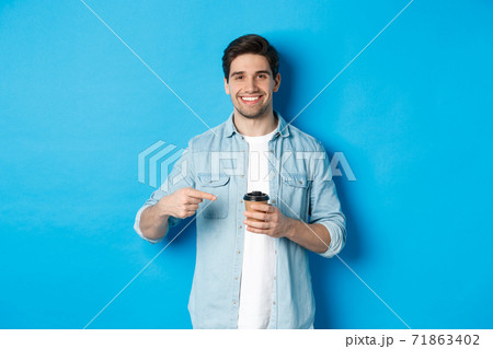 Smiling happy man pointing at paper cup with coffee, recommending cafe, standing over blue background Smiling happy man pointing at paper cup with coffee, recommending cafe, standing over blue background 71863402