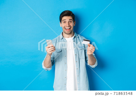 Amazed handsome man shopping online, holding mobile phone and credit card, smiling while paying for internet purchase, standing over blue background 71863548