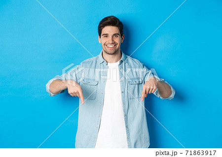 Handsome smiling adult man introduce product, pointing fingers down at promotion, standing against blue background 71863917
