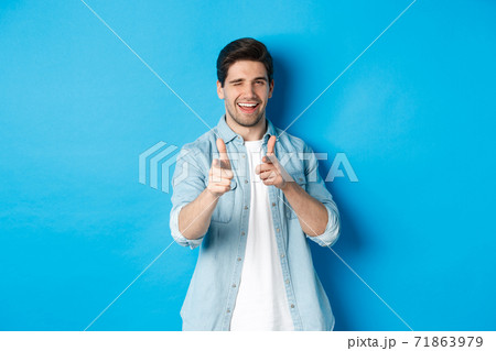 Cheeky handsome guy pointing fingers at you, winking flirty, standing in casual outfit against blue background 71863979