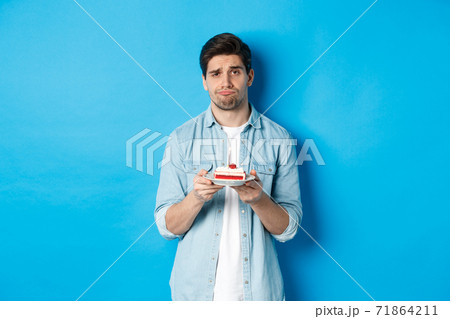 Upset and gloomy man holding birthday cake, sulking sad, standing against blue background Upset and gloomy man holding birthday cake, sulking sad, standing against blue background 71864211
