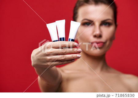 Beauty portrait of a young woman on red. The woman shows tubes with creams. Beauty portrait of a young woman on red. The woman shows tubes with creams. 71865830