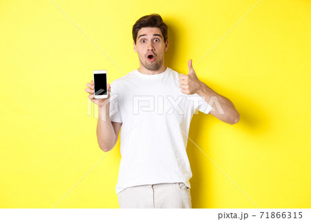 Impressed guy showing awesome smartphone app on screen and thumb-up, gasping amazed, standing against yellow background 71866315