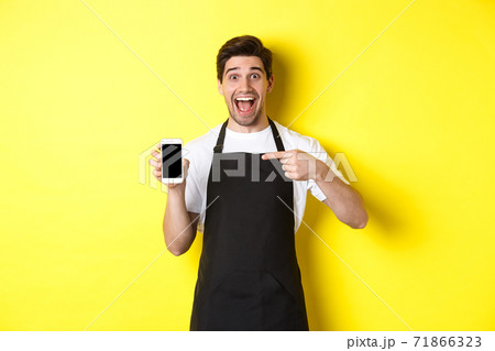Handsome barista in black apron pointing finger at mobile screen, showing app and smiling, standing over yellow background 71866323