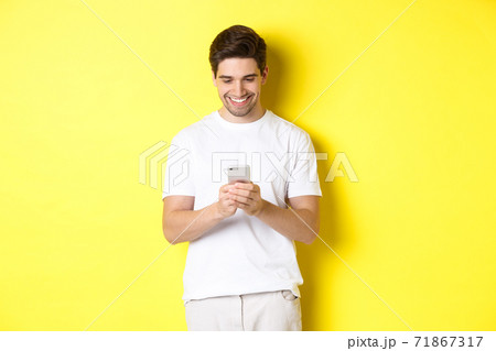 Young man reading text message on smartphone, looking at mobile phone screen and smiling, standing in white t-shirt against yellow background 71867317