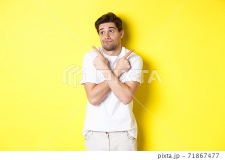 Indecisive man shrugging, pointing fingers sideways, troubled to choose, standing over yellow background 71867477