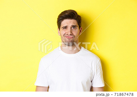 Close-up of dispelased young man in white t-shirt looking doubtful, grimacing unsatisfied, standing over yellow background 71867478