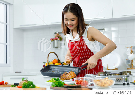 Asian woman chef in red apron holding hot steak with tong before serving in dish. Concept woman preparing meals at home. 71869474