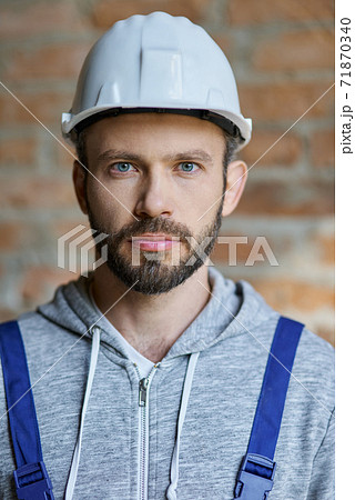 Close up portrait of confident attractive young male builder in hard hat looking at camera, posing on a brick wall background, working at construction site 71870340
