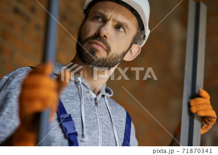 Close up shot of young male builder wearing hard hat looking focused, holding metal studs for drywall while working at construction site Close up shot of young male builder wearing hard hat looking focused, holding metal studs for drywall while working at construction site 71870341