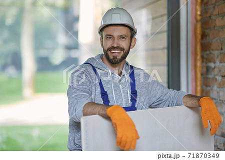 Portrait of handsome young male builder in hard hat looking positive, holding drywall while working at construction site 71870349