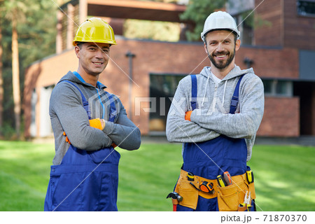 Experts in remodelling. Portrait of two handsome young male engineers in hard hats smiling at camera, posing outdoors with arms crossed while working on cottage construction Experts in remodelling. Portrait of two handsome young male engineers in hard hats smiling at camera, posing outdoors with arms crossed while working on cottage construction 71870370