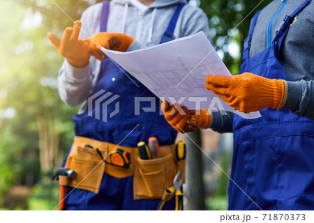 New project. Close up shot of two builders standing outdoors with an open blueprint discussing the cottage construction New project. Close up shot of two builders standing outdoors with an open blueprint discussing the cottage construction 71870373