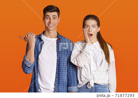 Surprised man smiles and points finger to free space, amazed female with open mouth presses hand to her cheek 71877293