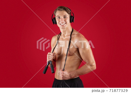 Sexy young sportsman with jumping rope wearing headphones, listening to music on red studio background Sexy young sportsman with jumping rope wearing headphones, listening to music on red studio background 71877329