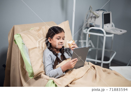 Positive little girl using an app on her digital device, while staying in a hospital bed Positive little girl using an app on her digital device, while staying in a hospital bed 71883594
