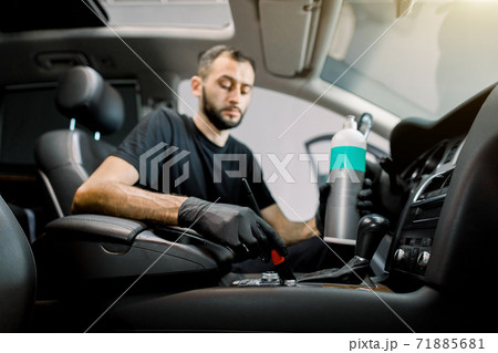Car interior plastic care, detailing concept. Portrait of young handsome male Caucasian auto service worker, cleaning car interior with soft brush and special professional care milk. Focus on the hand 71885681