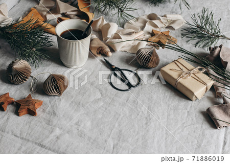 Christmas festive still life composition. Cup of coffee, paper Christmas ornaments and gift box. Velvet ribbons, pine tree branches with scissors. Linen table cloth background. Moody winter banner. Christmas festive still life composition. Cup of coffee, paper Christmas ornaments and gift box. Velvet ribbons, pine tree branches with scissors. Linen table cloth background. Moody winter banner. 71886019