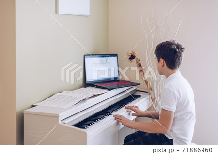 12 year old boy playing the white piano looking at the notes of his laptop. 71886960