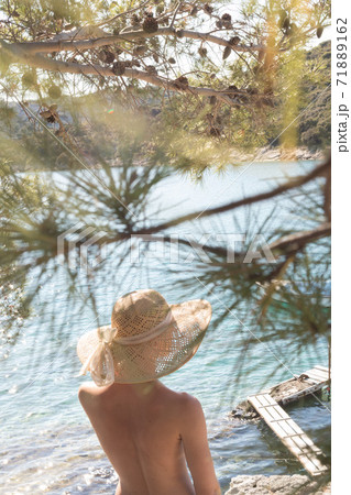 Rear view of topless beautiful woman wearing nothing but straw sun hat realaxing on wild coast of Adriatic sea on a beach in shade of pine tree. 71889162