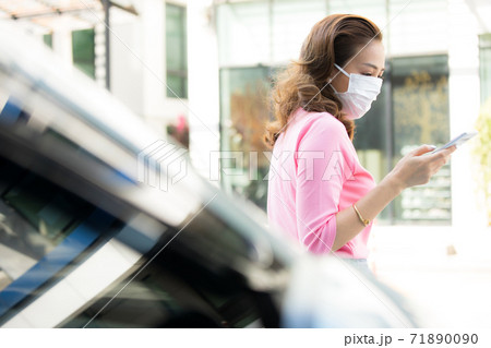自動車の前で立って携帯電話を使っているフェースマスク姿の若い女性 71890090