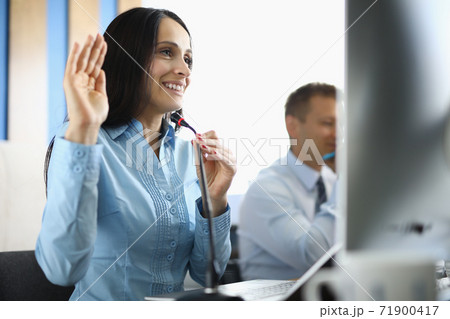 Businesswoman sits at computer with microphone smiling and raising her hand in a welcoming manner. 71900417