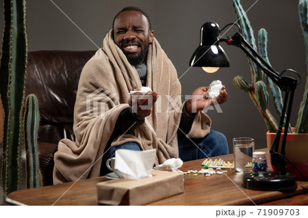 Young man wrapped in a plaid looks sick, ill, sneezing and coughing sitting at home indoors. 71909703