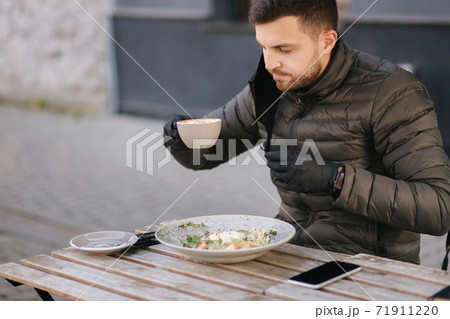 Man in black gloves hold cutlery. Quarantine Cafe concept. Food outdoors in protective gloves. Fresh vegan salad with shrimps. Man pulled off protective mask for drinking cappuccino Man in black gloves hold cutlery. Quarantine Cafe concept. Food outdoors in protective gloves. Fresh vegan salad with shrimps. Man pulled off protective mask for drinking cappuccino 71911220