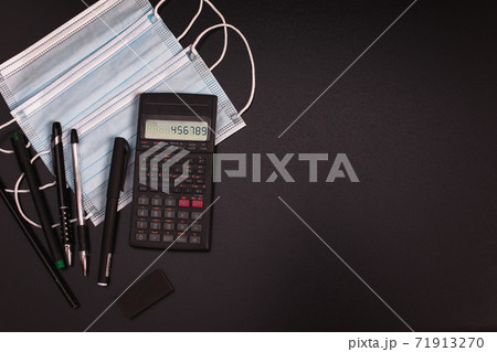 Calculator, pens, protective face mask on black backgroung. Remote Work from Home Office during Corona Virus Pandemic. 71913270