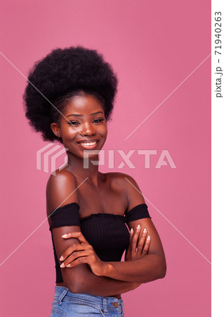 Beautiful African American girl with gorgeous afro hairstyle standing smiling with arms folded in black top and denim jeans isolated on pink background 71940263