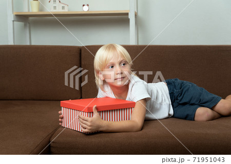 Child lies on the couch with gift box in his hands. Blond boy holding bright box in his hands 71951043