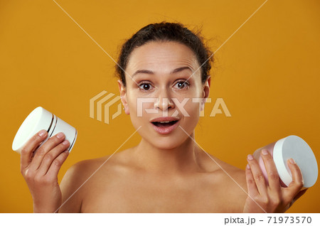 Beautiful african woman looks surprised at camera while holding jars of creams isolated on yellow Beautiful african woman looks surprised at camera while holding jars of creams isolated on yellow 71973570