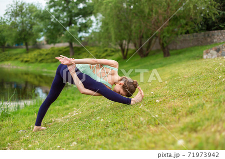 Slender young brunette yogi performs challenging yoga exercises on the green grass in the summer against the backdrop of nature 71973942