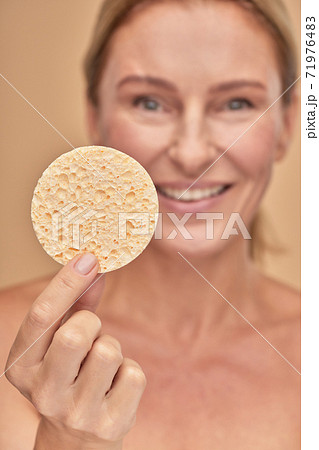 Vertical shot of a charming mature female holding cleaning makeup sponge while standing isolated over beige background in studio 71976483